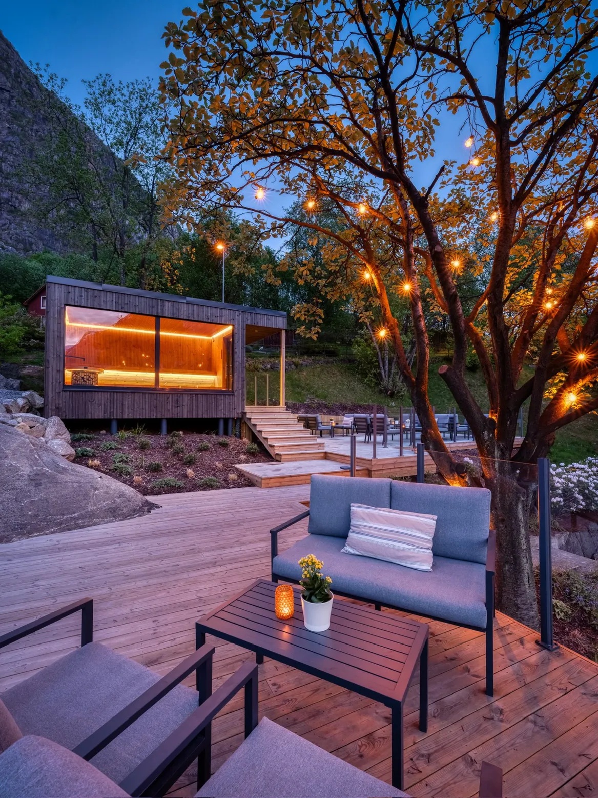 Widok na strefę z sauną. NAsadzenia na skarpie ściółkowane korą. Oświetlone ażurowe drzewo.