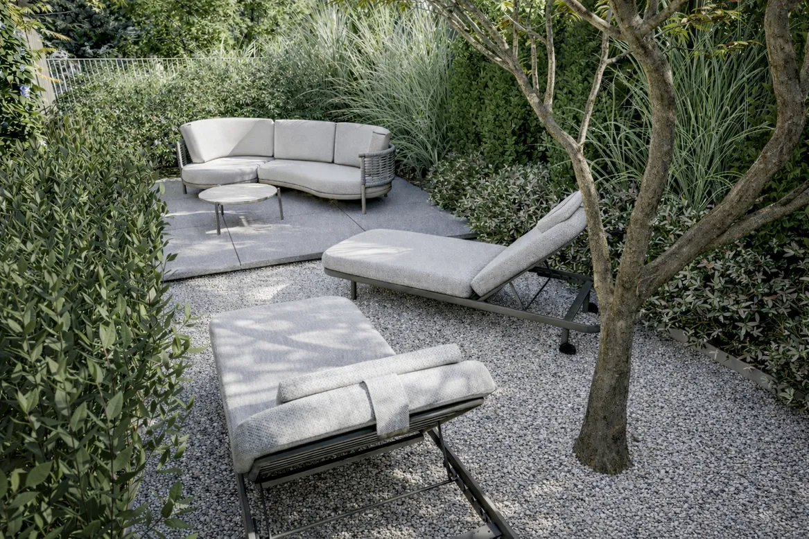 Widok na zaciszną strefę relaksacyjną otoczoną gęstą zielenią. Szare poduszki mebli komponują się z kolorem żwiru i elementami betonowymi.
