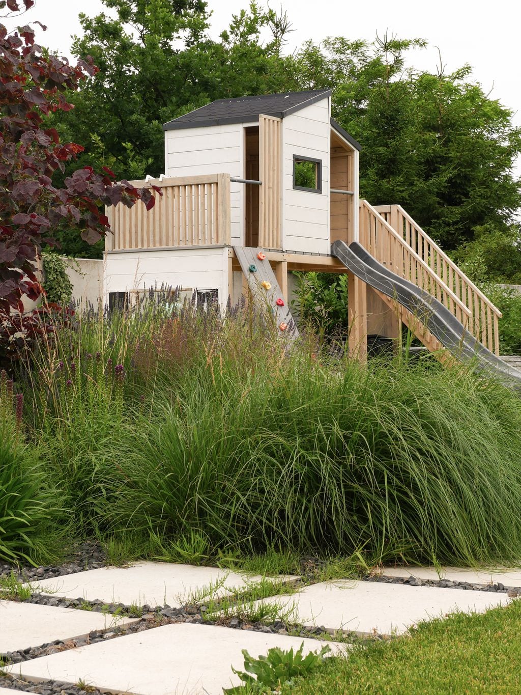 Domek zabaw dla dzieci z drugiej strony. Ścieżka z jasnych płyt betonowych oddzielonych żwirem. Trawy, byliny. 